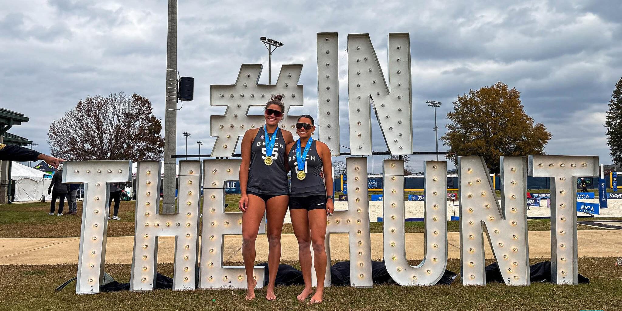 Long Beach State winning beach VB pair standing in front of the "In The Hunt" sign